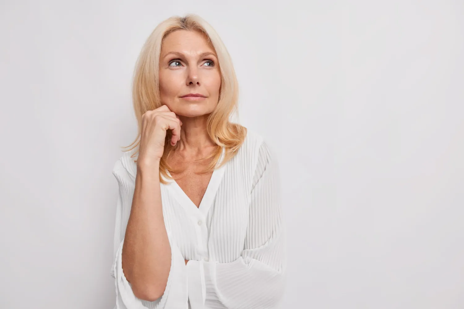 thoughtful-blonde-middle-aged-woman-ponders-something-keeps-hand-near-face-has-healthy-skin-minimal-makeup-makes-choice-wears-white-blouse-poses-indoor-blank-copy-space-your-promotion